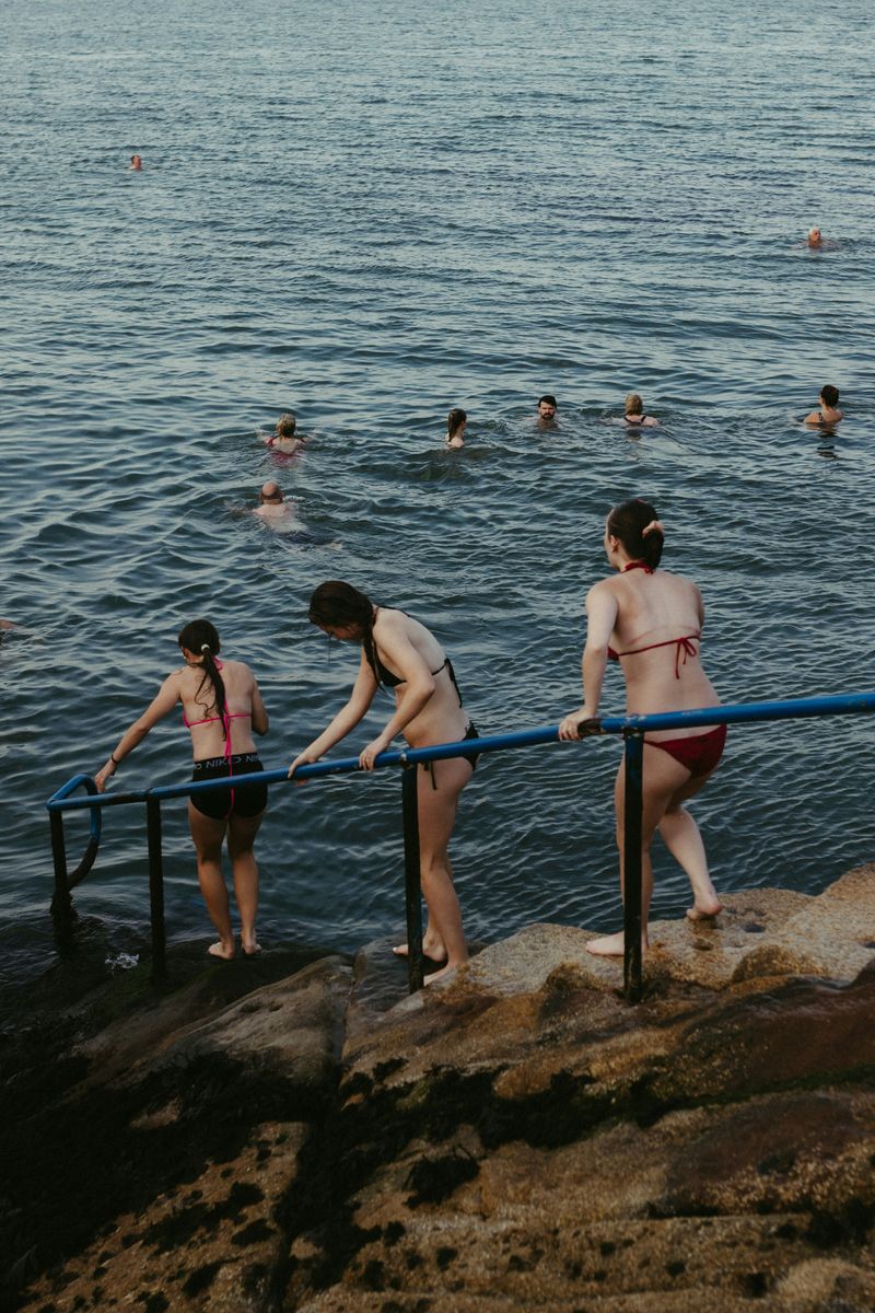 Mehrere Menschen in Badekleidung steigen über ein blaues Geländer von Felsen ins Meer, weitere schwimmen bereits