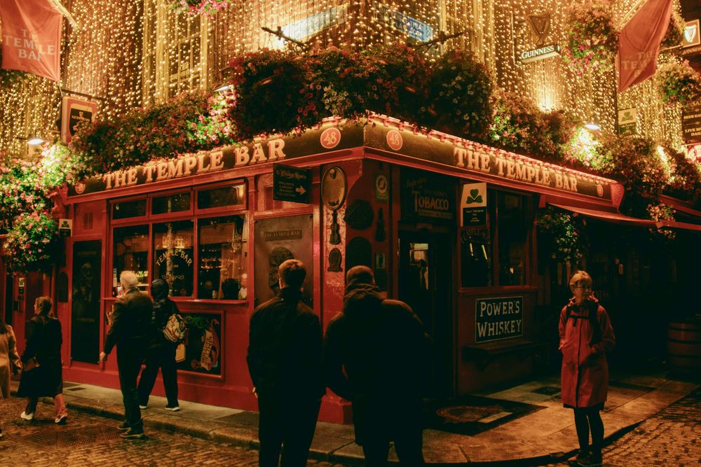 Die rote Fassade des Pubs The Temple Bar in Dublin bei Nacht, überzogen von Lichterketten, Passanten davor