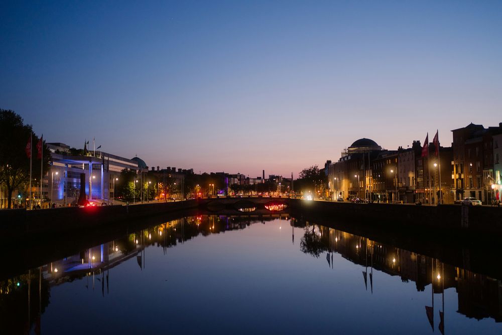 Die Liffey in Dublin bei Abenddämmerung, spiegelglattes Wasser, beleuchtete Kais und die Kuppel der Four Courts