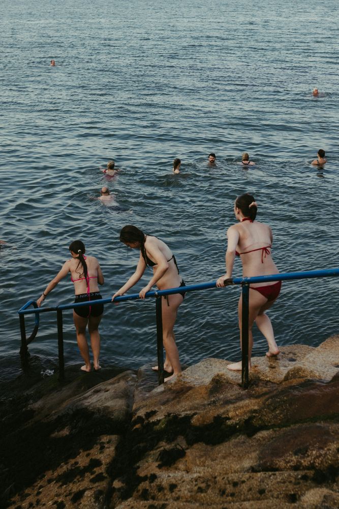 Mehrere Menschen in Badekleidung steigen über ein blaues Geländer von Felsen ins Meer, weitere schwimmen bereits