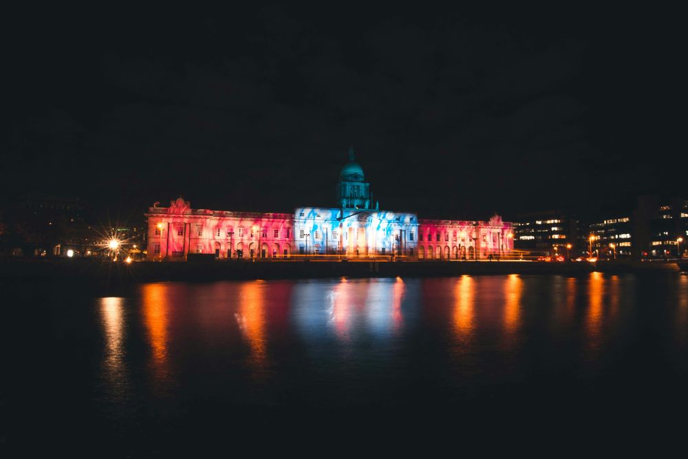 Ein historisches Gebäude in Dublin bei Nacht farbig angestrahlt, gespiegelt im Wasser der Liffey